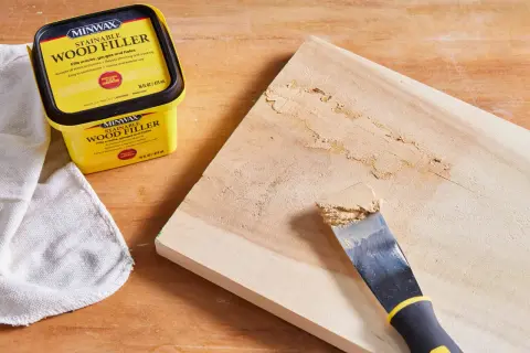 Un pot de mastic à bois Minwax est à côté d'une planche de bois où un couteau à mastic applique le produit pour reboucher bois.