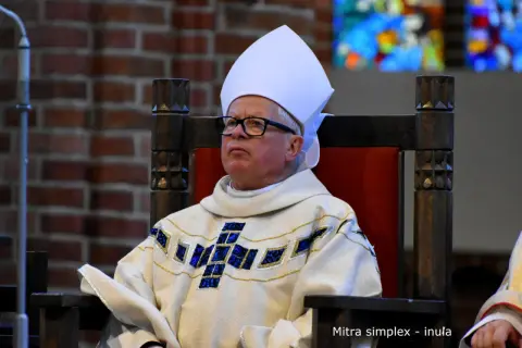 Biskup w białej mitrze i ozdobnej szacie z krzyżem, siedzi na drewnianym tronie.