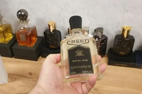 Kolekcja perfum Creed, w tym Creed Royal Oud, prezentowana na drewnianym blacie. W dłoni trzymana jest butelka Creed Royal Oud.