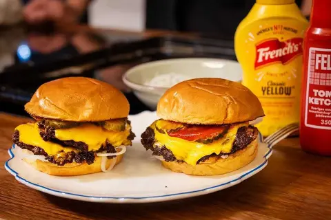 Två saftiga smashed burgers med smält ost, pickles och lök. Bredvid står flaskor med senap och ketchup.
