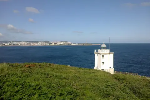 El faro de Mera se alza blanco sobre un promontorio verde, vigilando la costa y el mar azul.