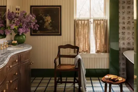Ambiance de salle de bain rustique avec un lavabo en marbre, des lilas violets, un tableau ancien et une chaise en bois.