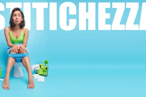 Ragazza con espressione preoccupata seduta sul water, circondata da prodotti per l'igiene. La parola "STITICHEZZA" campeggia in alto. Forse le patate non sono la soluzione.