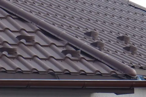 Brązowy dach z dachówki ceramicznej, widoczne elementy systemu rynnowego i wentylacyjnego. Jak zrobić zakończenie gąsiora?