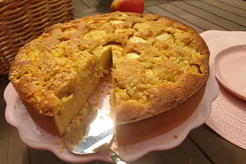 Ein goldbrauner Apfelkuchen ohne Zucker, bestreut mit Nüssen, wird auf einem rosa Teller präsentiert. Daneben liegt ein roter Apfel.