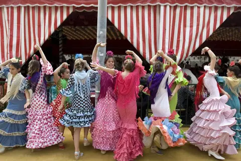 Niñas con trajes de volantes y lunares, aprendiendo a bailar sevillanas con arte bajo una carpa a rayas.
