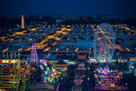 Nocne, rozświetlone atrakcjami wesołego miasteczka, gdzie trwa **feria de abril**. Widać diabelski młyn, kolejki górskie i tłumy ludzi.