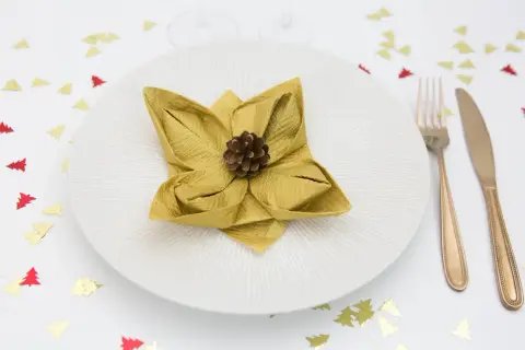 Une serviette dorée pliée en forme de fleur de lotus, ornée d'une pomme de pin, sur une assiette blanche. Confettis sapins rouges et dorés sur la table.