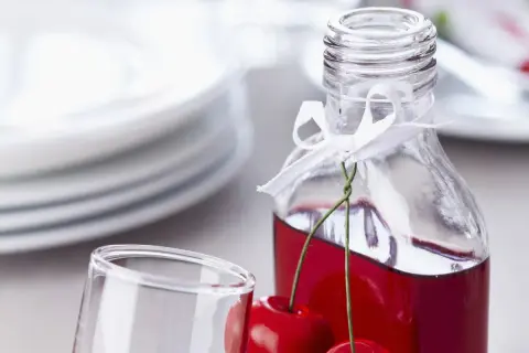 Elegancka butelka z wiśniowym alkoholem, ozdobiona wstążką i świeżymi wiśniami, obok kieliszka i stosu talerzy.