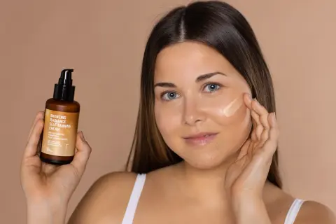 Mujer aplica bronceador freshly en su mejilla. Sostiene un frasco de crema autobronceadora.