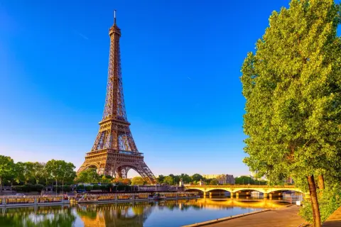 Eiffeltornet i Paris, med sina tre våningar, speglar sig i Seine. En grönskande trädtopp skymtar till höger.