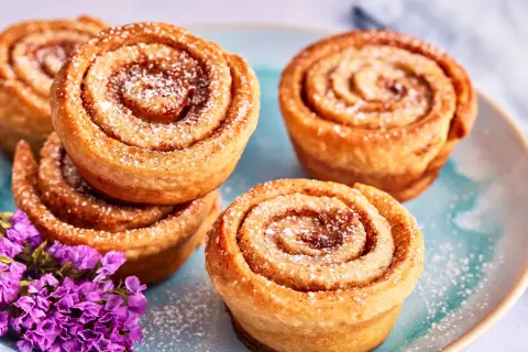 Zimtschnecken Muffins, bestäubt mit Puderzucker, liegen auf einem blauen Teller neben lila Blumen.