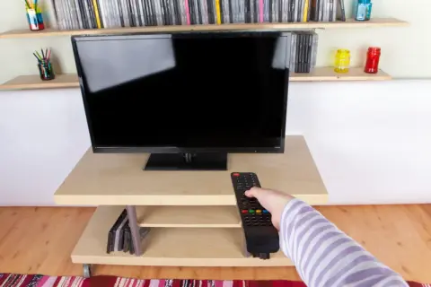 Ręka z pilotem sięga do telewizora naprzeciwko okna. Na półkach stoją płyty i kolorowe słoiki.