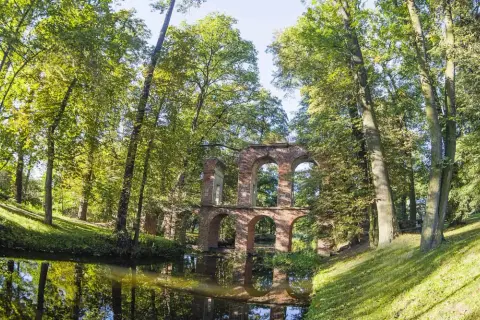 Kamienne ruiny mostu w Arkadii Nieborów odbijają się w spokojnej wodzie, otoczone bujną zielenią drzew.
