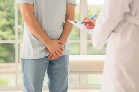 Mężczyzna w jasnej koszulce i jeansach stoi przed lekarzem w białym kitlu, który notuje coś na kartce. To wizyta kontrolna, być może związane z badaniem krwi na prostatę.