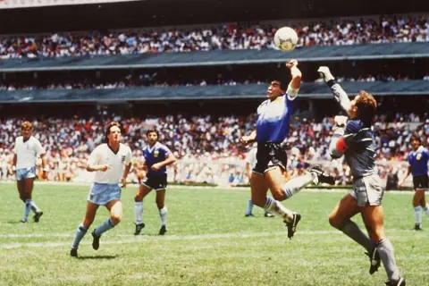 Diego Maradona, w słynnym meczu, używa "ręka boga", by zdobyć bramkę. Piłkarze w białych strojach patrzą z niedowierzaniem.