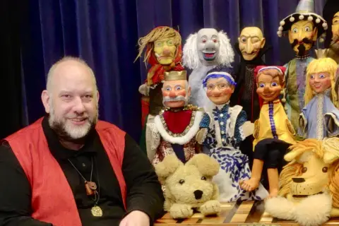 Ein Mann lächelt neben einer bunten Truppe von Handpuppen, die für den Puppenpavillon Bensberg bereitstehen.