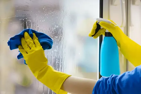 Manos con guantes amarillos limpian cristales muy sucios con un paño azul y spray.