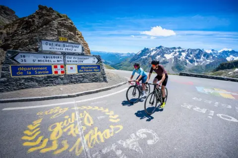 Deux cyclistes gravissent le col du Galibier, un paysage alpin spectaculaire sous un ciel bleu éclatant.