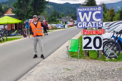 Pracownik w kamizelce odblaskowej kieruje ruchem na parkingu siwa polana. W tle góry i budynki.