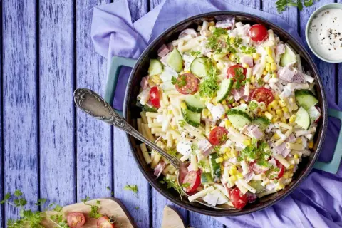 Ein köstlicher nudelsalat mit schinken, garniert mit Kirschtomaten, Gurkenscheiben und Mais. Perfekt für ein Sommerfest!