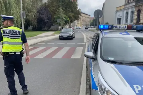 Policjant w kamizelce odblaskowej i radiowóz z włączonym sygnałem świetlnym stoją przy przejściu dla pieszych. Na radiowozie widoczny jest znak d6.