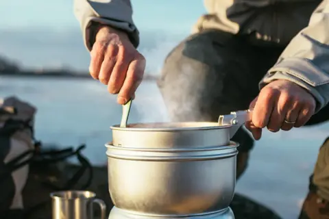 En person lagar mat på Trangia utomhus. Ånga stiger från kastrullen medan maten rörs om.