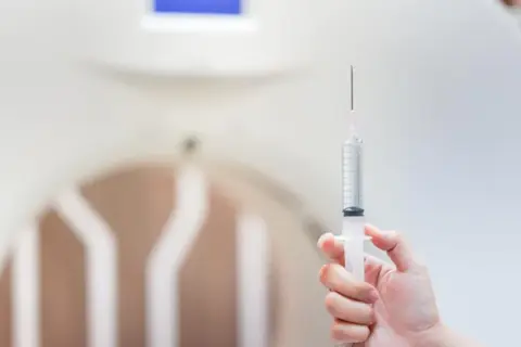 Dłoń trzyma strzykawkę z płynem. W tle widać fragment aparatu do rezonansu magnetycznego, który pomaga uwidocznić szczegóły, tworząc kontrast.