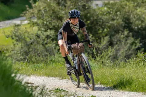 Kobieta z tatuażami na rowerze, ciesząca się kolarstwem gravelowym na piaszczystej drodze wśród zieleni.