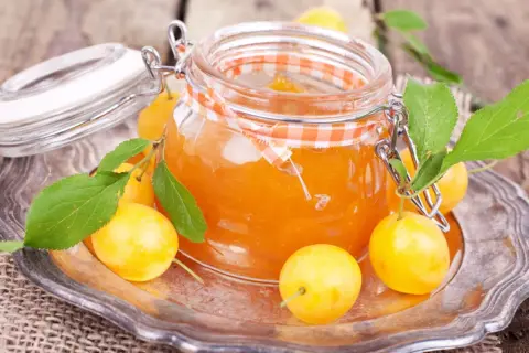 Ein Glas mit leuchtend orangefarbener, raffinierter Mirabellenmarmelade, umgeben von frischen Mirabellen und grünen Blättern auf einem silbernen Teller.