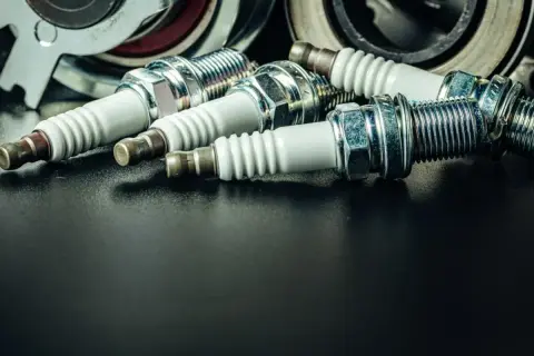 Kilka świec zapłonowych, niektóre z widocznym nalotem, leżą obok części mechanicznych.