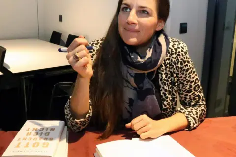Esther Maria Magnis hält einen Stift und blickt nachdenklich. Auf dem Tisch liegt ihr Buch "Gott braucht dich nicht".