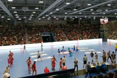 Handballspiel in der Gummersbach Schwalbe Arena. Zuschauer füllen die Ränge, Spieler auf dem Feld, eine Maskottchen-Figur und Cheerleader sind zu sehen.