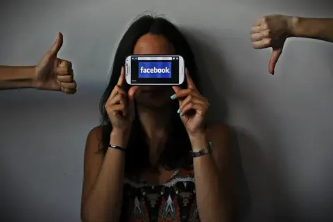 Una persona oculta su rostro tras un móvil con Facebook. A los lados, pulgares arriba y abajo, reflejando el impacto de las redes sociales y la autoestima.