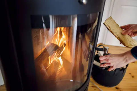 Ein Kaminofen mit brennendem Holz. Eine Hand hält ein Holzscheit, bereit zum Nachlegen. Bald muss der alte Kaminofen entsorgt werden.