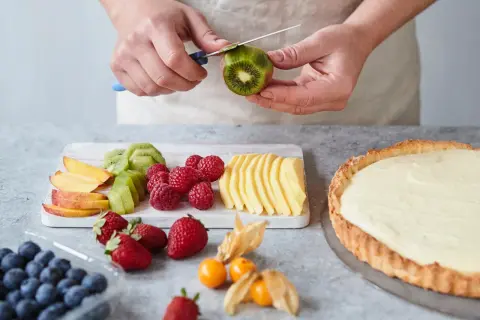 Hände schälen Kiwi für ein fruchtiges Tarte Rezept. Daneben liegen Beeren, Pfirsichspalten und Apfelscheiben.