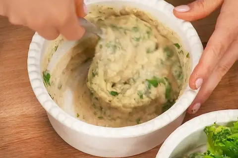 En hand rör i en skål med krämiga refried beans och färska örter.