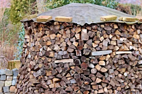 Ein großer Stapel Brennholz, der mit einer Plane und Ziegeln abgedeckt ist. Die Holzscheite sind ordentlich aufgeschichtet, bereit für den Winter. Die Baustahlmatte dient als stabile Unterlage.