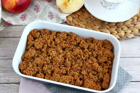 En härlig glutenfri frukost med äppelcrisp i en vit form, äpplen och en kopp te i bakgrunden.