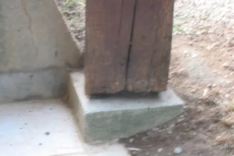 Un poteau bois pourri à la base repose sur un socle en béton. La dégradation du bois est visible.