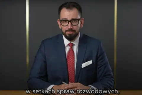 Mężczyzna w garniturze i okularach siedzi przy biurku, trzymając długopis.
