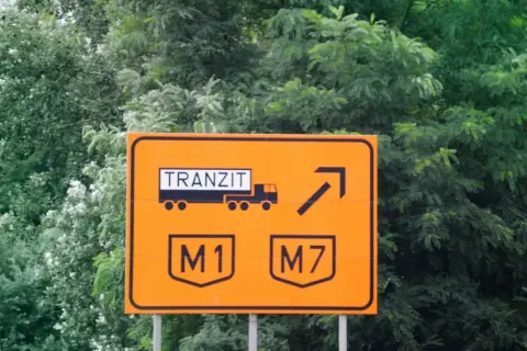 Pomarańczowy znak drogowy z napisem "TRANZIT" i symbolem ciężarówki, wskazujący zjazd na autostrady M1 i M7. Winieta węgry.