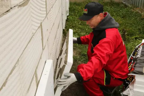 Pracownik w czerwonym stroju montuje płyty styropianu na ścianie z bloczków. To kluczowy etap prac, który decyduje o tym, ile styropianu na strop betonowy będzie potrzebne.