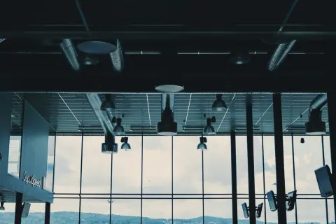 Ventilationsrör och lampor i taket, med en stor fönstervägg som visar en molnig himmel och en grönskande kulle. Kanske är det en plats där man kontrollerar radon nivåer.