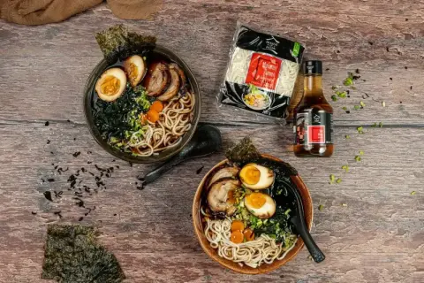 Dwa miski aromatycznego ramen z jajkiem, mięsem i wodorostami. Obok makaron ramen i sos.