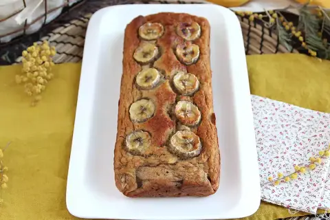 Delizioso banana bread light, decorato con fette di banana, su un piatto bianco, con fiori di mimosa sullo sfondo.