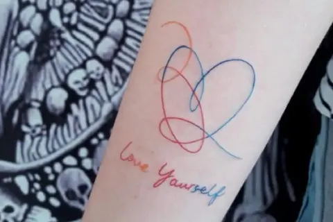 Tatuajes de corazón y "Love Yourself" en el brazo, un símbolo de fortaleza y amor propio.