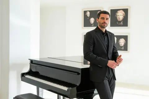 Uomo in elegante outfit teatro, con completo nero, accanto a un pianoforte a coda.