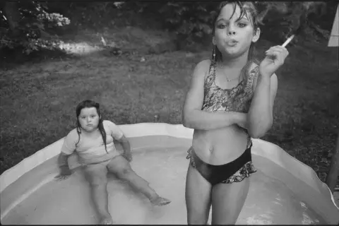Deux jeunes filles dans une piscine gonflable, l'une fumant une cigarette, dans le style de Mary Ellen Mark.