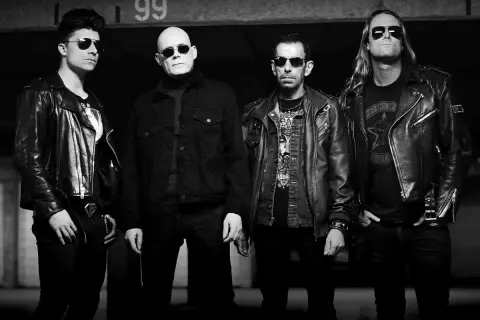 Czterech mężczyzn w skórzanych kurtkach, w tym jeden z napisem "Sisters of Mercy" na koszulce, pozuje do zdjęcia.